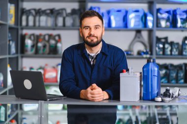 Adam araba parçaları atölyesinde çalışıyor, dizüstü bilgisayarı, aküsü ve motor yağıyla ayakta duruyor, kaliteli otomotiv parçaları sunuyor.