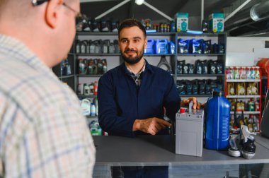Otomotiv işçisi kasada müşteriyle konuşuyor. Araba aküsünü ve motor yağını oto yedek parça mağazasında gösteriyor.