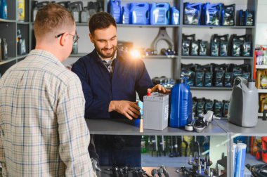 Otomobil işçisi, yeni bir araba aküsü ve motor yağıyla müşteriye yardım ediyor, oto yedek parçalarıyla tezgahta duruyor.