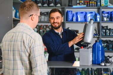 Otomotiv parçaları mağazasındaki müşteriye motor yağı ve araba aküsünü gösteren satıcı, hizmet ve uzmanlık sunar