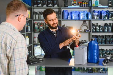 Bir oto yedek parça dükkanının tezgahındaki müşteriye anahtarı gösteren tamirci, ayrıca araba aküsü ve motor yağı da sunuyor.