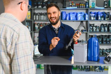 Oto yedek parça mağazası temsilcisi gülümsüyor, araba parçası tutuyor, müşteriye otomotiv ürünleri ve araba aküsü hakkında tavsiye veriyor.