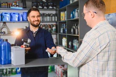 Otomobil servis teknisyeni elinde bir ingiliz anahtarı tutuyor ve erkek bir müşteriyle pil ve yağ hakkında konuşuyor.