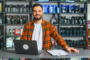 Otomotiv tezgahında dizüstü bilgisayar kullanan ve kameraya bakan genç bir erkek satıcı. Raflar araba yağıyla dolu.