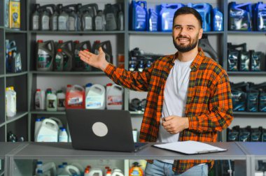 Gülümseyen genç bir adam, motor yağı ve bakım sıvılarıyla dolu bir tamirhanede dizüstü bilgisayar ve pano kullanarak duruyor.