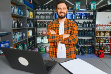 Genç bir erkek satıcı, motor yağı ve araç gereçleri olan bir mağazada dizüstü bilgisayarını iş için kullanıyor.