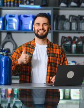 Gülümseyen oto tamircisi araba servisi atölyesinde duruyor, başparmak işareti veriyor ve motor yağı olan bir dizüstü bilgisayar kullanıyor.