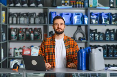 Bir oto yedek parça mağazasında çalışan, dizüstü bilgisayarı, barkod tarayıcısı ve motor yağı kutularıyla tezgahta duran bir adam.