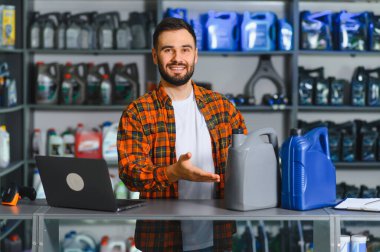Gülümseyen adam otomobil parçaları veya kayganlaştırıcı dükkanında motor yağı kutularını sunuyor, rafların önünde duruyor.