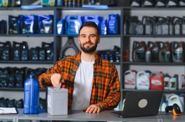 Araba yedek parça dükkanındaki tezgahta gülümseyen satıcı, araba aküsünü raflarda yağ ve kayganlaştırıcı ile satılmaya hazır tutuyor.