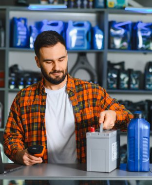 Oto yedek parça mağazasında çalışan genç adam, araba aküsünü tarıyor ve arka planda yağ şişeleriyle envanter yönetiyor.