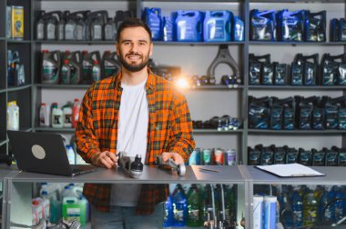 Bir oto yedek parça mağazasında çalışan, gülümseyen sakallı bir adam, yedek araç parçaları tutuyor, raflarda yağlar ve yağlar var.