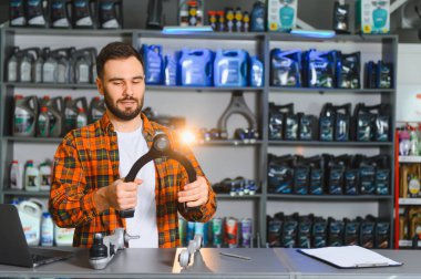 Bir oto yedek parça dükkanındaki adam satış için bir kontrol kolunu tutuyor ve inceliyor, arkasında da kayganlaştırıcı ve araba parçaları var.