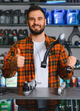 Gülümseyen adam oto yedek parça mağazasında araba süspansiyon kontrol kolunu sunuyor, ayrıca baş parmak işareti gösteriyor.