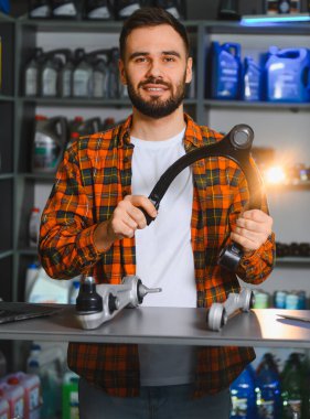 Ekose gömlekli sakallı tamirci, galeride top eklemli bir kontrol kolu ve oto yedek parça tezgahının üzerinde çalılıklar tutuyor.