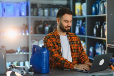 Envanter ve satışları internet üzerinden yöneten bir erkek mağaza müdürü, bir oto yedek parça dükkanında yağ şişeli bir dizüstü bilgisayar kullanıyor.