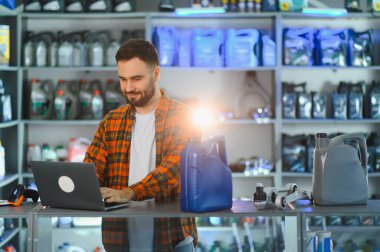 Erkek mağaza çalışanı dizüstü bilgisayarda envanter yönetimi ve sipariş işleme yapıyor, bir oto yedek parça dükkanında tezgahın önünde duruyor.