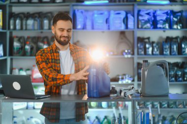 Kareli gömlekli ve sakallı bir adam, bir oto yedek parça mağazasındaki raflardan bir şişe motor yağı seçerken bir müşteriye yardım ediyor.