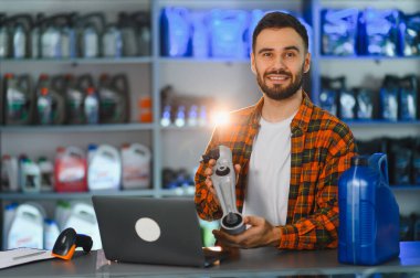 Araba parçaları dükkanında çalışan satıcı, süspansiyon parçası tutuyor, dizüstü bilgisayar kullanıyor, raflarda motor yağı var.