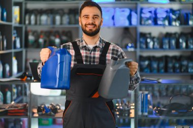 Bir oto yedek parça mağazasında çalışan gülümseyen tamirci, elinde iki şişe motor yağı tutuyor, araç bakımı için seçenekler sunuyor.