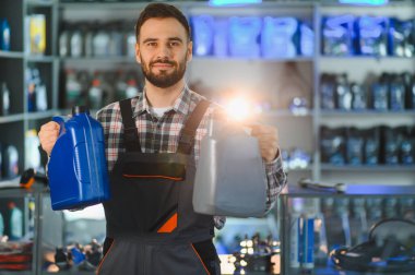 Gülümseyen tamirci oto yedek parça dükkanında duruyor, iki şişe motor yağı sunuyor ve kameraya bakıyor.