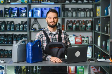 Otomobil parçaları mağazası çalışanı tezgahta duruyor, bir dizüstü bilgisayar üzerinde çalışıyor, motor yağı kapları ve ürünlerle dolu raflarla