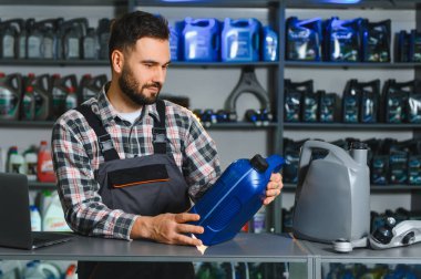 Tezgahta bir şişe motor yağını inceleyen iş elbiseli bir adam. Etrafı araba yağlarıyla ve raflardaki parçalarla çevrili.