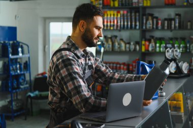 Ekose gömlekli ve tulumlu bir satıcı oto yedek parça reyonunda bir şişe motor yağını bilgisayarla tarıyor.