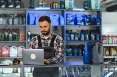 Araba bakım atölyesinde dizüstü bilgisayar kullanan oto servis teknisyeni, motor yağı ve yedek parçalarla çevrili.