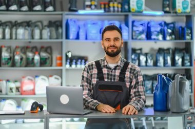 Araba servisi çalışanı tulum ve ekose gömlek giyer, bir araba parçacıda dizüstü bilgisayarla ve motor yağıyla tezgaha gülümser.