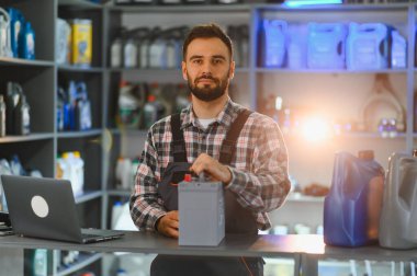 Araba yedek parça dükkanında profesyonel satış danışmanı elinde araba aküsü, elinde dizüstü bilgisayarla tezgahın arkasında duruyor.