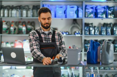 Bir oto yedek parça dükkanında dizüstü bilgisayarı, araba aküsü ve yağ bidonlarıyla çalışan oto servis teknisyeni.