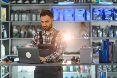 Erkek işçi tulum giyiyor ve ekose gömlek giyiyor. Bir oto yedek parça mağazasında dizüstü bilgisayar kullanıyor. Arkaplanda petrol ve araba aküleri olan raflar
