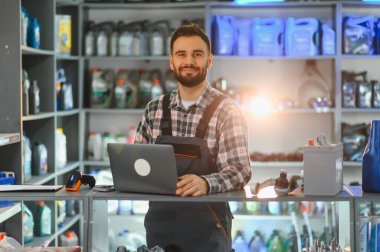 Tezgahta çalışan, arkasında otomobil parçaları ve ürünleri sergilenen dizüstü bilgisayarlı bir adam.