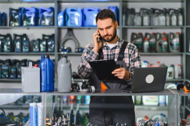 Oto yedek parça mağazasında çalışan servis teknisyeni, telefon siparişi alıyor, panoyu tutuyor ve dizüstü bilgisayardaki envanteri kontrol ediyor.