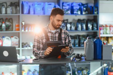 Tulumlu bir adam stokları inceliyor, araba yağı şişeleriyle tezgahta duruyor ve perakende oto yedek parça dükkanında pil satıyor.