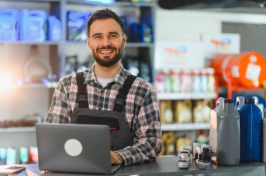 Araba parçaları ve yağ şişeleri olan oto tamircisinin kasasında dizüstü bilgisayar kullanan mutlu bir tamirci.
