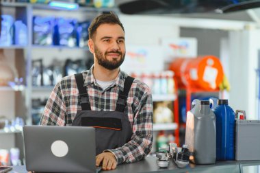 Tulumlu ve ekose gömlekli sakallı tamirci, masa başında otomobil parçaları ve dizüstü bilgisayarla gülümsüyor, envanter ve online siparişleri yönetiyor.
