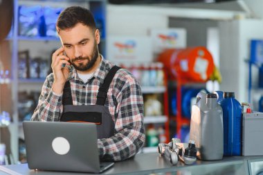 Tezgahta çalışan oto yedek parça mağazası çalışanı, bir telefon görüşmesi yapıyor ve müşteri hizmetleri ve envanter için dizüstü bilgisayar kullanıyor.