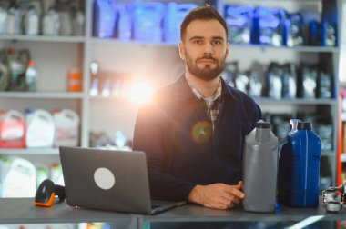 Bir oto yedek parça mağazasında tezgahın arkasında elinde motor yağı şişeleriyle duran bir satıcı. Dizüstü bilgisayar ve barkod tarayıcısı masada.