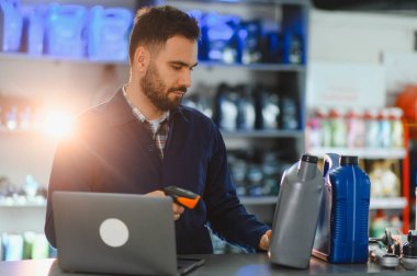 Araba servisi uzmanı bir oto yedek parça dükkanında motor yağı şişelerini tarıyor, bir barkod okuyucusu ve dizüstü bilgisayarla envanteri yönetiyor.