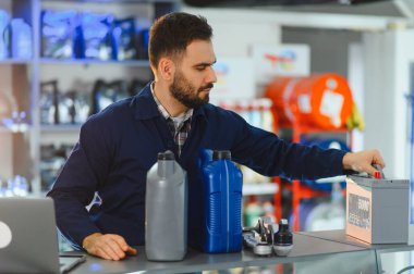 Servis çalışanı garajdaki bir masada yeni bir araba aküsü, motor yağı ve otomobil parçaları ile ilgileniyor.