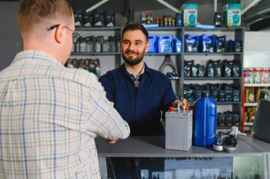 Gülümseyen tamirci ya da satıcı bir oto yedek parça mağazasında araba aküsü ve motor yağı sunan müşteriye tezgahta yardım ediyor.