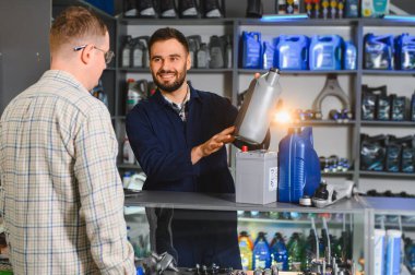 Otomobil parçaları işçisi, erkek müşteriye yardım ediyor, bir şişe motor yağı tutuyor ve otomotiv sıvısı ve pil seçimini gösteriyor
