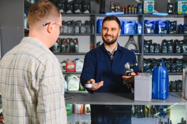 Gülümseyen bir tamirci bir araba deposunda araba aküsü, yağ ve parçalarla müşteriye yardım ediyor.