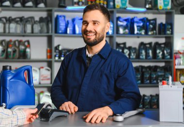 Gülümseyen tamirci bir oto yedek parça mağazasında kredi kartı ödeme terminalini kullanarak müşteriye akü ve motor yağı satıyor.