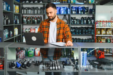 Bir oto yedek parça mağazasında dizüstü bilgisayarda çalışan bir dükkan sahibi, envanter ve iş operasyonlarını yönetiyor. Birçok araba parçası görünür.