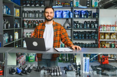 Çeşitli otomotiv malzemeleri ve parçalarıyla çevrili işlek bir oto yedek parça dükkanında envanter ve satışları yöneten gülümseyen adam.