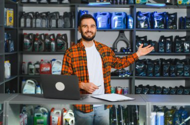 Gülümseyen, dizüstü bilgisayarla bir oto yedek parça mağazasında çalışan, motor yağı ve araba bakım malzemeleriyle dolu rafları olan bir adam.