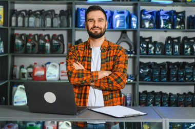 Erkek tezgahtar ya da dükkan sahibi çapraz kollarla ayakta duruyor, perakende mağazasında araç yağı ve aksesuar satarken kameraya gülümsüyor.
